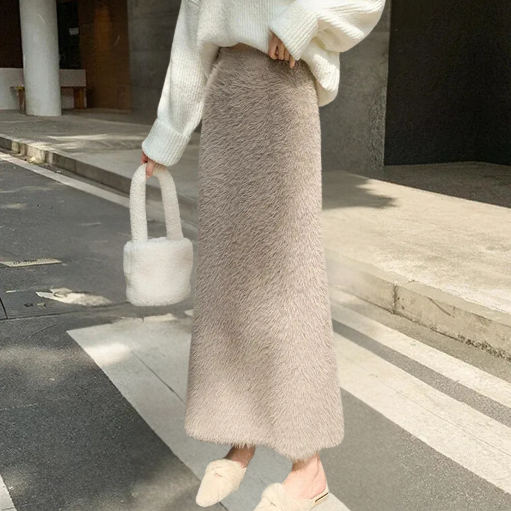 Vrouw in witte trui en lange beige pluizige rok, draagt een witte pluchen handtas en beige pantoffels, modieuze herfstoutfit op straat.